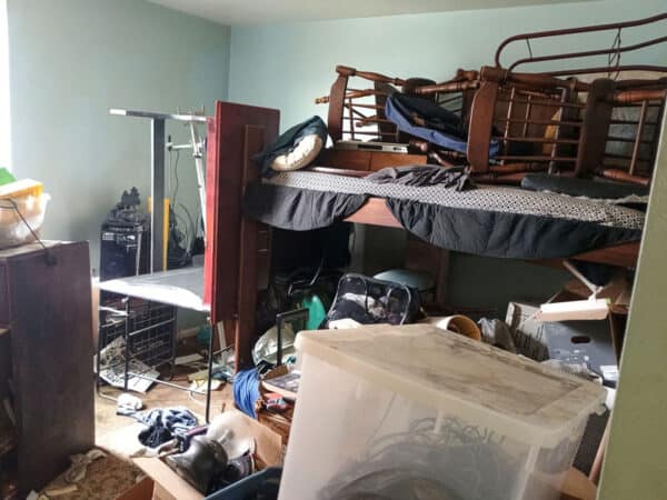 Messy bedroom with stacked furniture, clothes, and miscellaneous items.