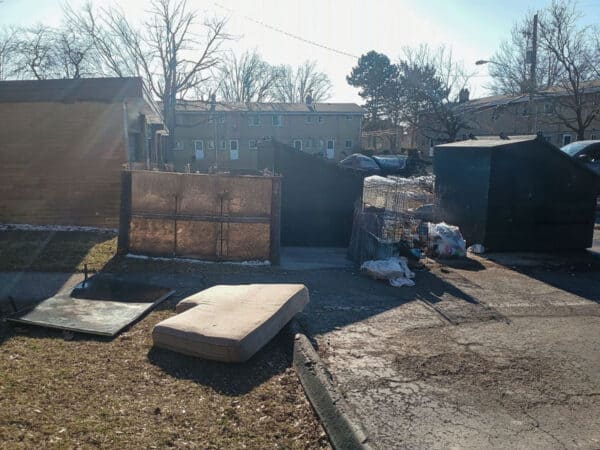 Outdoor scene with discarded mattress, debris, and storage sheds in a residential area.
