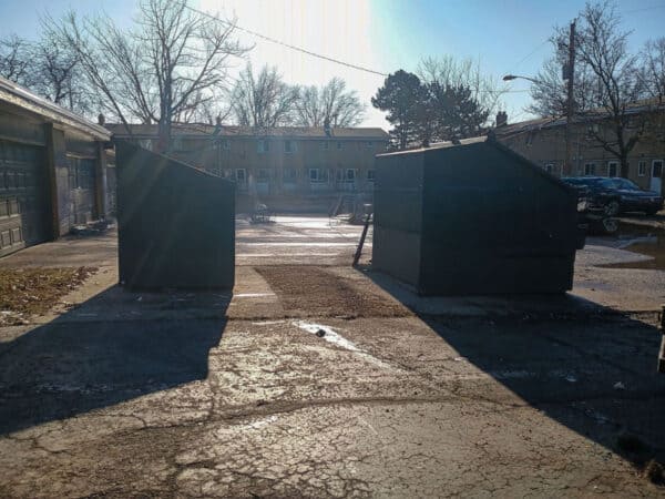 Two dark green storage sheds in an outdoor lot with shadows and trees in the background.