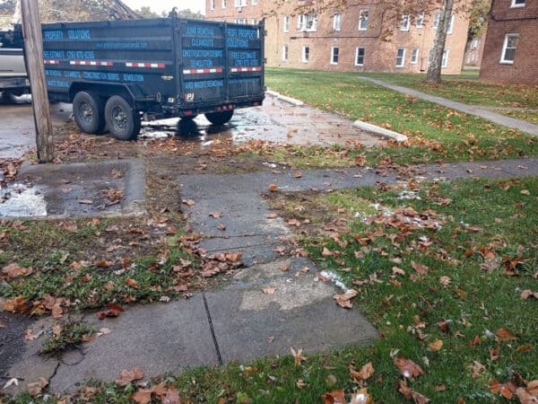 Large black dumpster on a residential street with fallen leaves and wet pavement.