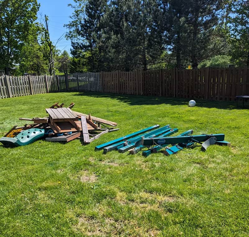 Disorganized backyard with scattered wooden and plastic debris on green grass.